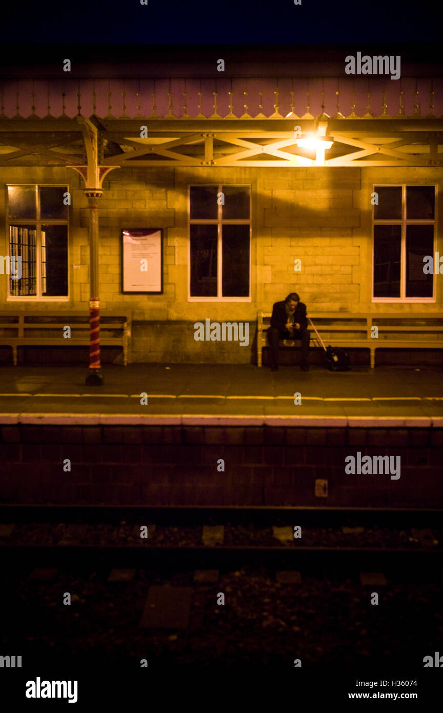 La figure solitaire d'un homme assis sur un banc dans une gare au Royaume-Uni. La nuit tombe et les lampes au sodium éclairent la scène avec la lueur orange familière et quelque peu étrange. L'homme a peut-être raté atrain ou le train pourrait être en retard car il a l'air d'utiliser un téléphone portable éventuellement pour faire savoir à ses proches quand il sera à la maison car il peut être bloqué pendant un certain temps. Banque D'Images