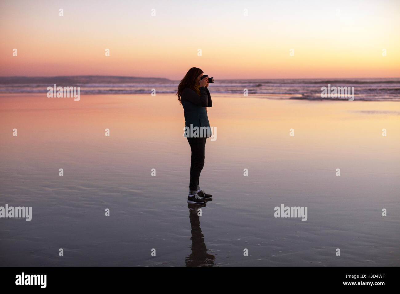 Vue de côté femme photographiant tout en se tenant à la plage pendant le coucher du soleil Banque D'Images