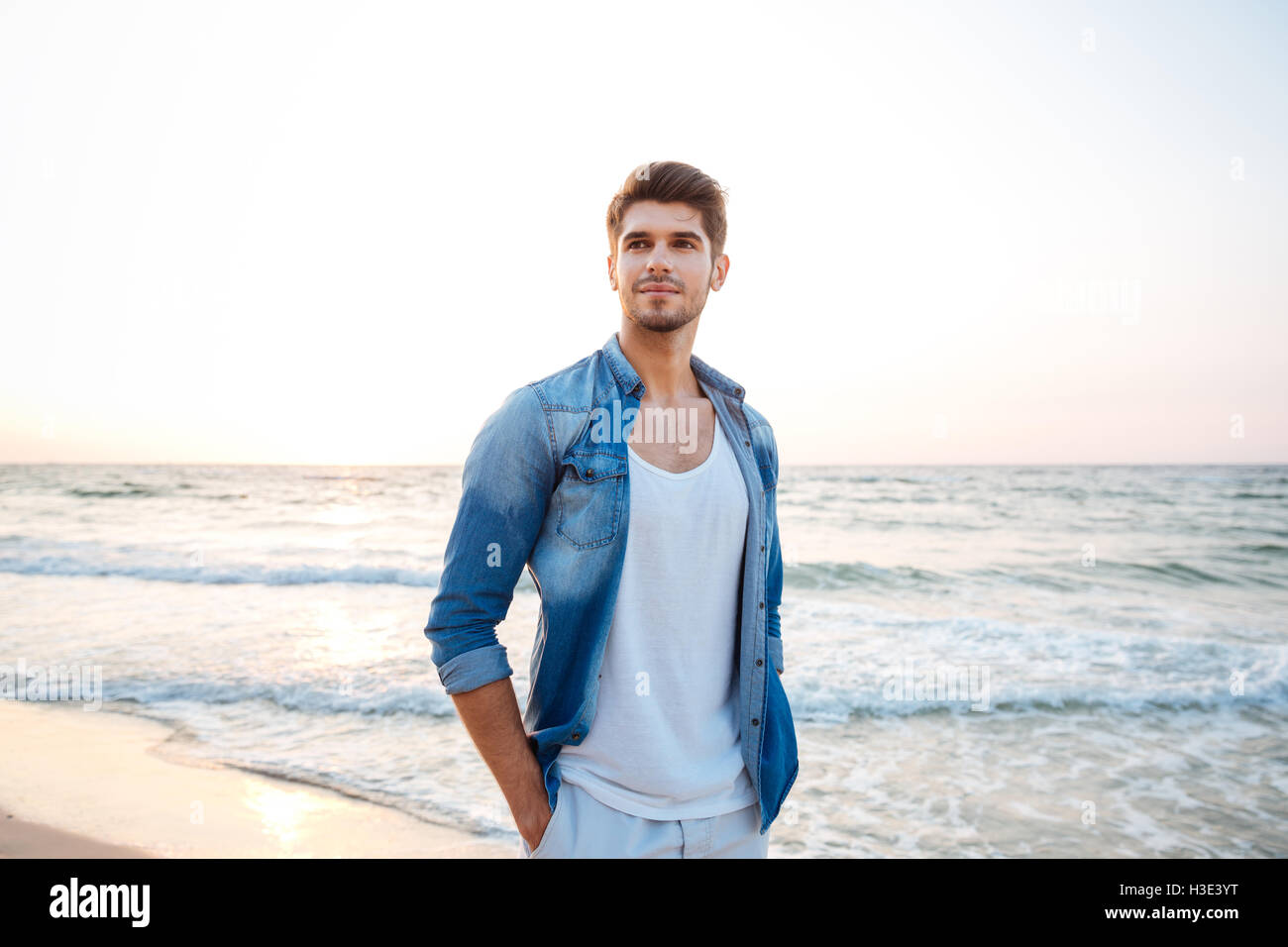 Beau jeune homme en jeans shirt debout sur la plage Banque D'Images