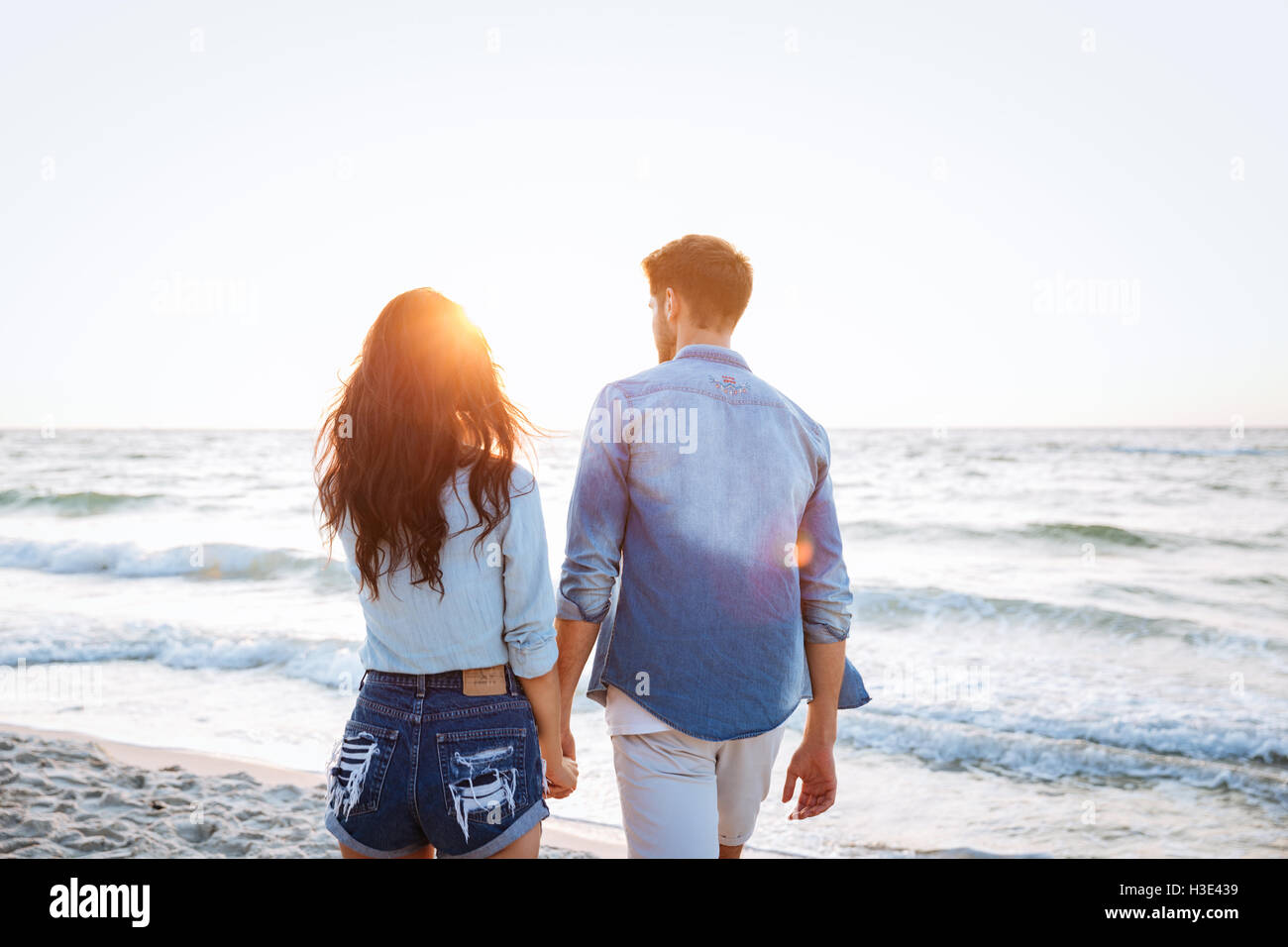 Vue arrière du jeune couple en train de marcher au lever du soleil sur la plage Banque D'Images