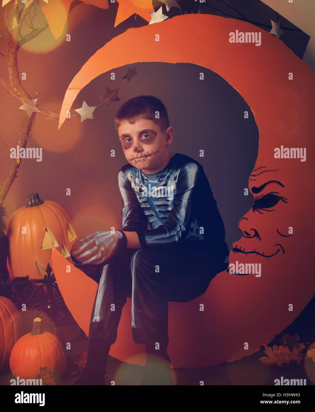 Un garçon est vêtu d'un costume de squelette Halloween et assis sur une lune orange avec un visage et des étoiles pour un portrait de nuit. Banque D'Images