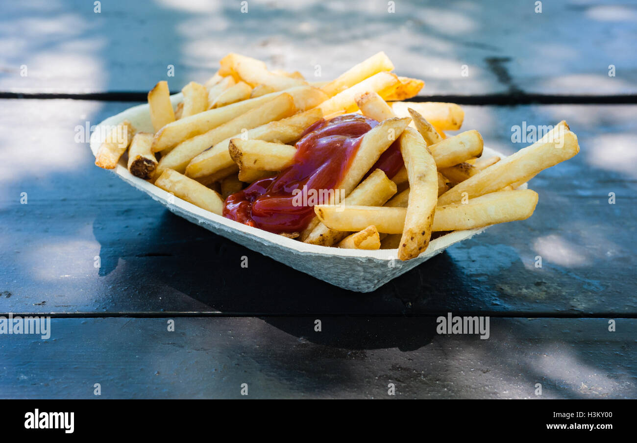 Des frites avec du ketchup contenant du papier sur bois table en lumière et ombre partielle à l'extérieur, vu de côté. Banque D'Images