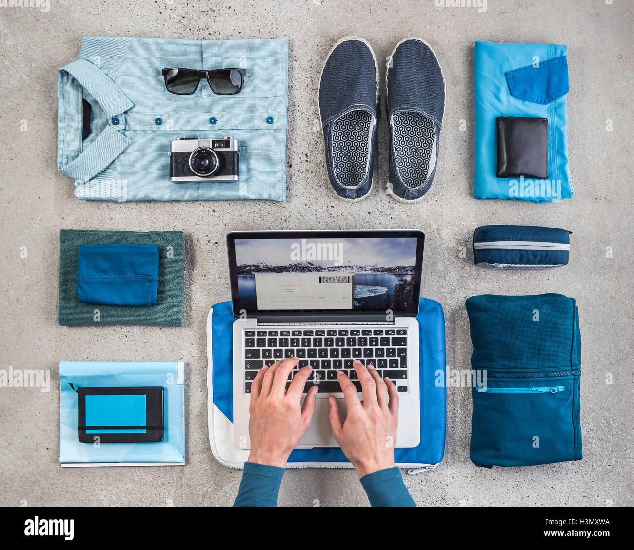 Vue de dessus de man's hands typing on laptop entouré par les éléments d'emballage, avec chemise bleu rétro, appareil photo, ordinateur portable et sac de lavage Banque D'Images