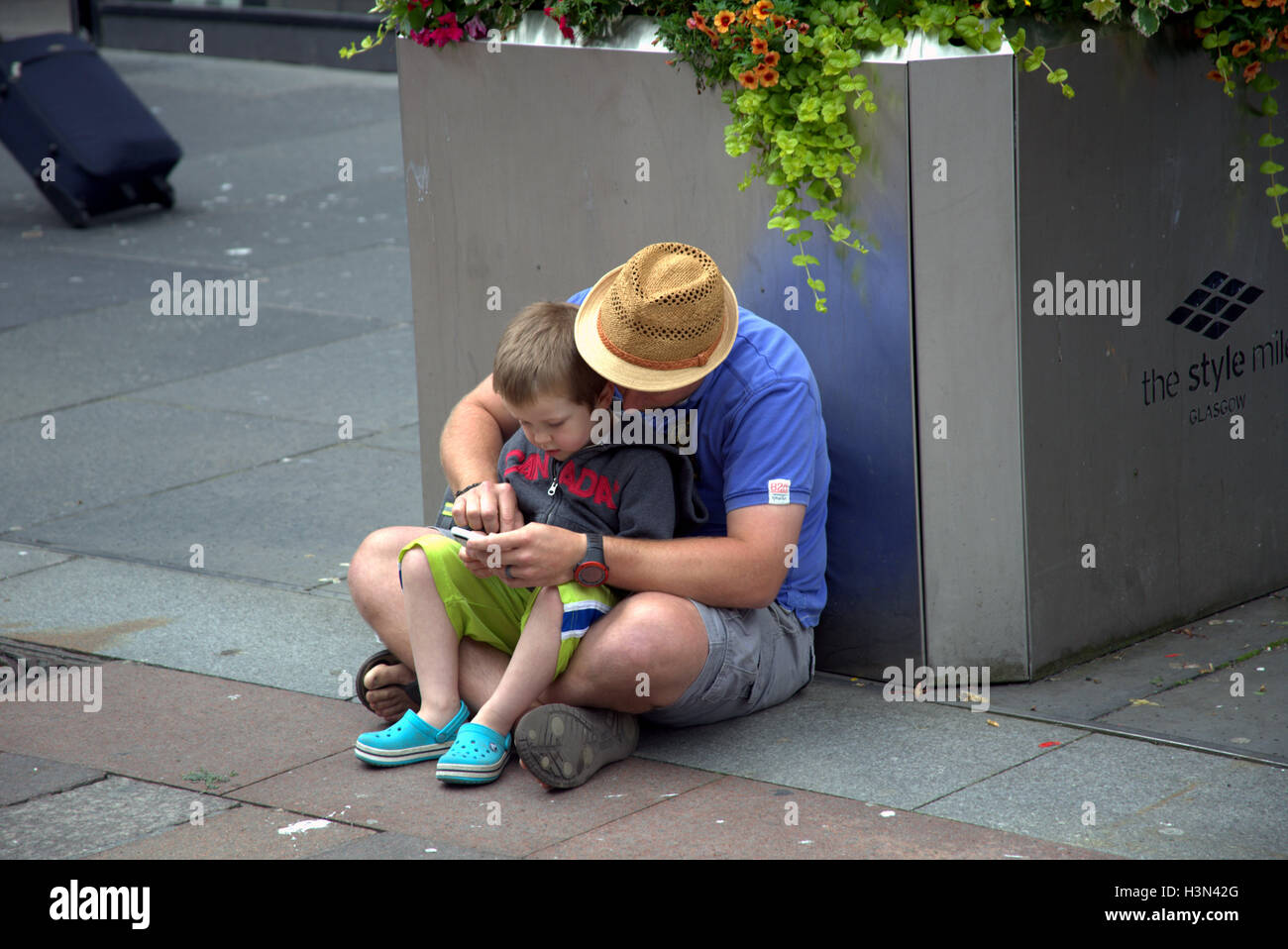 Scènes de rue de Glasgow père et fils utiliser mobile phone ensemble tout en restant assis sur le sol Banque D'Images