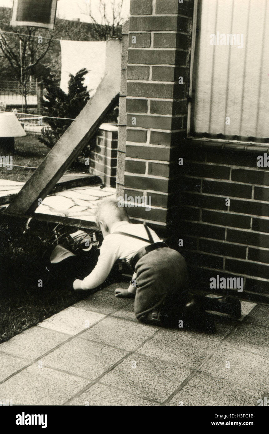 Allemagne - circa 1960 : petit garçon joue à quatre pattes sur le sol, mis à tuile, près de la chambre Banque D'Images