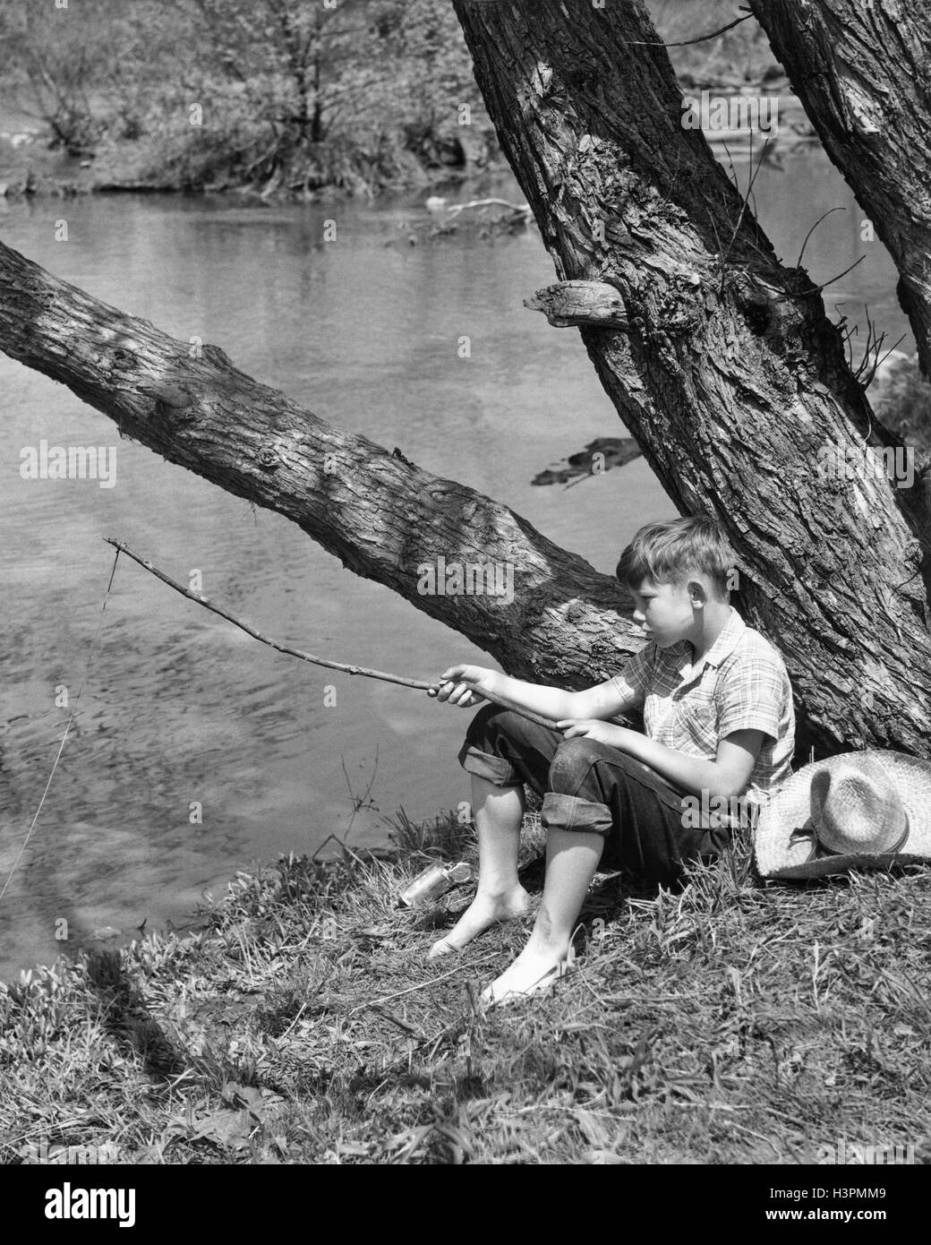 1940 BAREFOOT BOY SITTING UNDER TREE PAR FILIÈRE PÊCHE AVEC TWIG POLE Banque D'Images