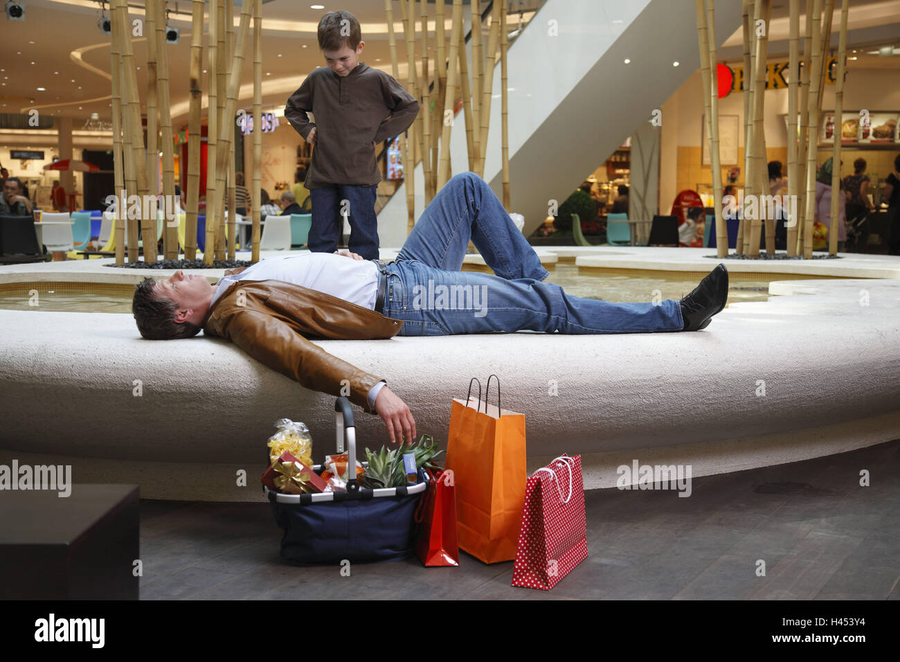 Centre Commercial, homme, se coucher, échappements, marge bien, garçon, impatiemment, stand, sur l'économie, au commerce, de la consommation, centre commercial, atrium, des boutiques, magasins, puits, fontaine, père, enfant, fils, personne, client, passant, exigeant, difficile, le contact oculaire, de reproche, résolument, se reposer, se détendre, fatigue, épuisement, rupture, l'attente, l'ennui, l'impatience, observer, faire des achats, shopping, sacs, sacs, sacs de transport, l'achat, à l'intérieur, Banque D'Images
