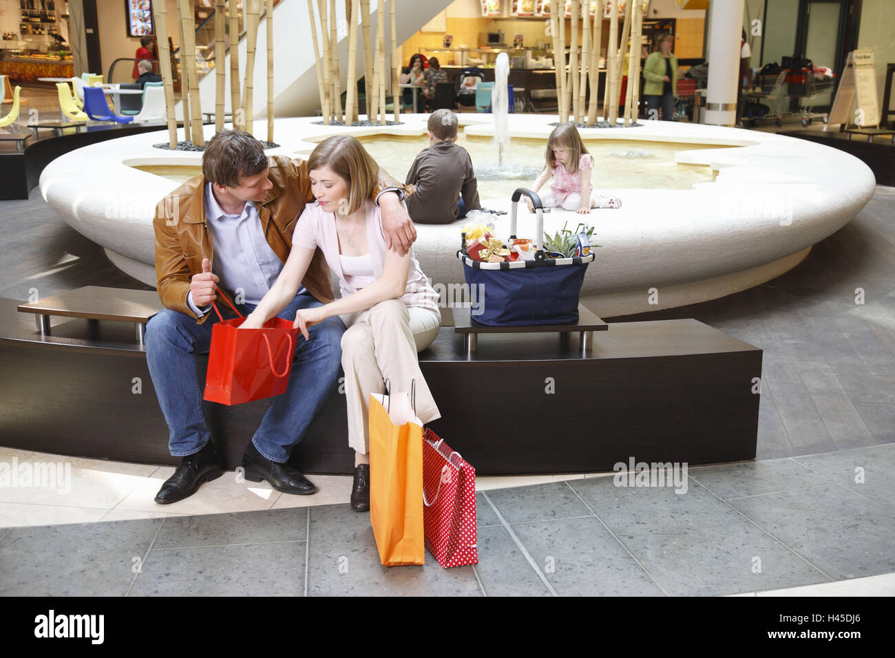Centre Commercial, famille, couple, satisfait, s'asseoir, de regarder l'achat, les puits, les enfants, jouer, l'économie, du commerce, de la consommation, centre commercial, atrium, magasins, boutiques, fontaine, l'écrou, le père, l'arrière-plan, garçon, fille, personne, client, passant, repos, faire des achats, shopping, sacs, sacs, sacs, à l'intérieur, Banque D'Images