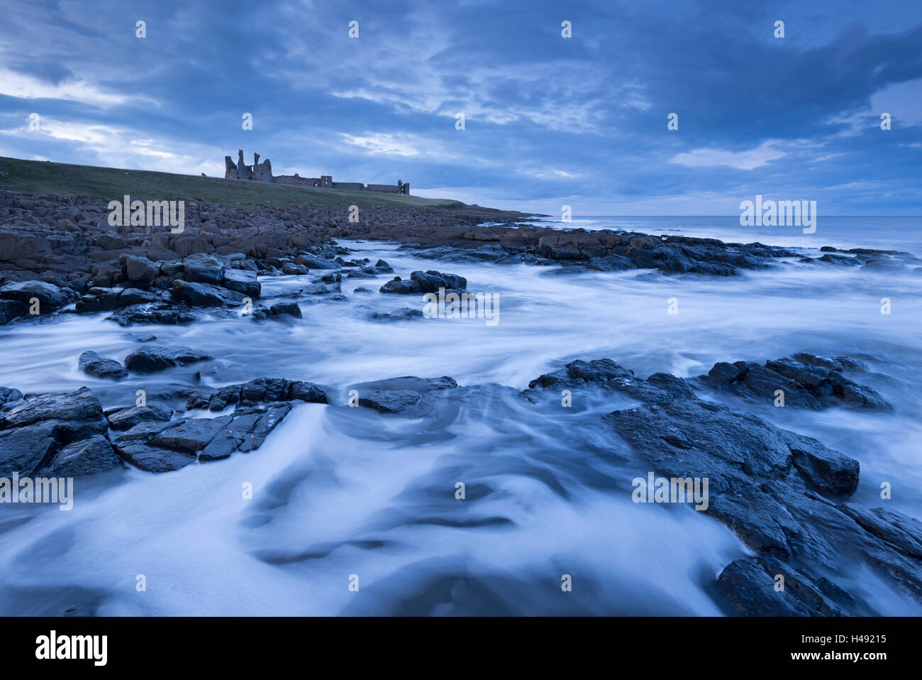 Vagues tourbillonnent autour les rivages rocheux ci-dessous château de Dunstanburgh au crépuscule, Northumberland, Angleterre. Printemps (mars) 2014. Banque D'Images