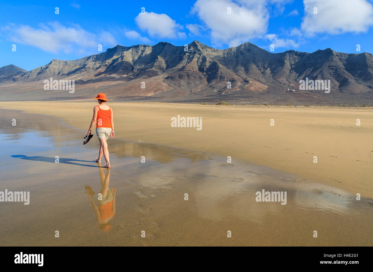 Jeune femme en chemise rouge et touristiques hat marche sur du sable doré humide plage de Cofete, Fuerteventura, Îles Canaries, Espagne Banque D'Images