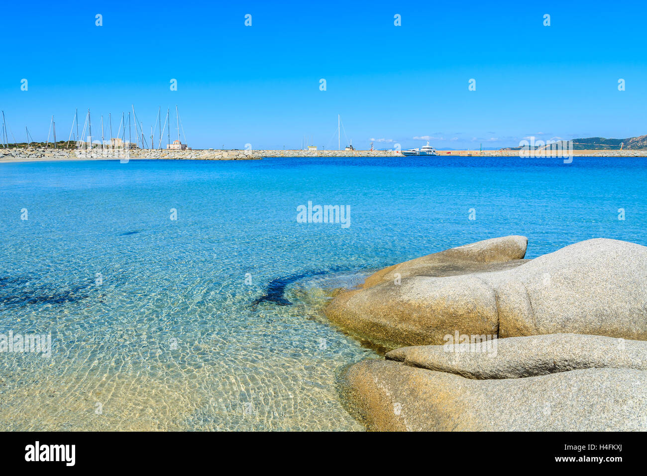 Des pierres dans l'eau de mer sur la plage de Spiaggia del Riso et port avec bateaux en arrière-plan, péninsule de Villasimius, Sardaigne, île, Italie Banque D'Images