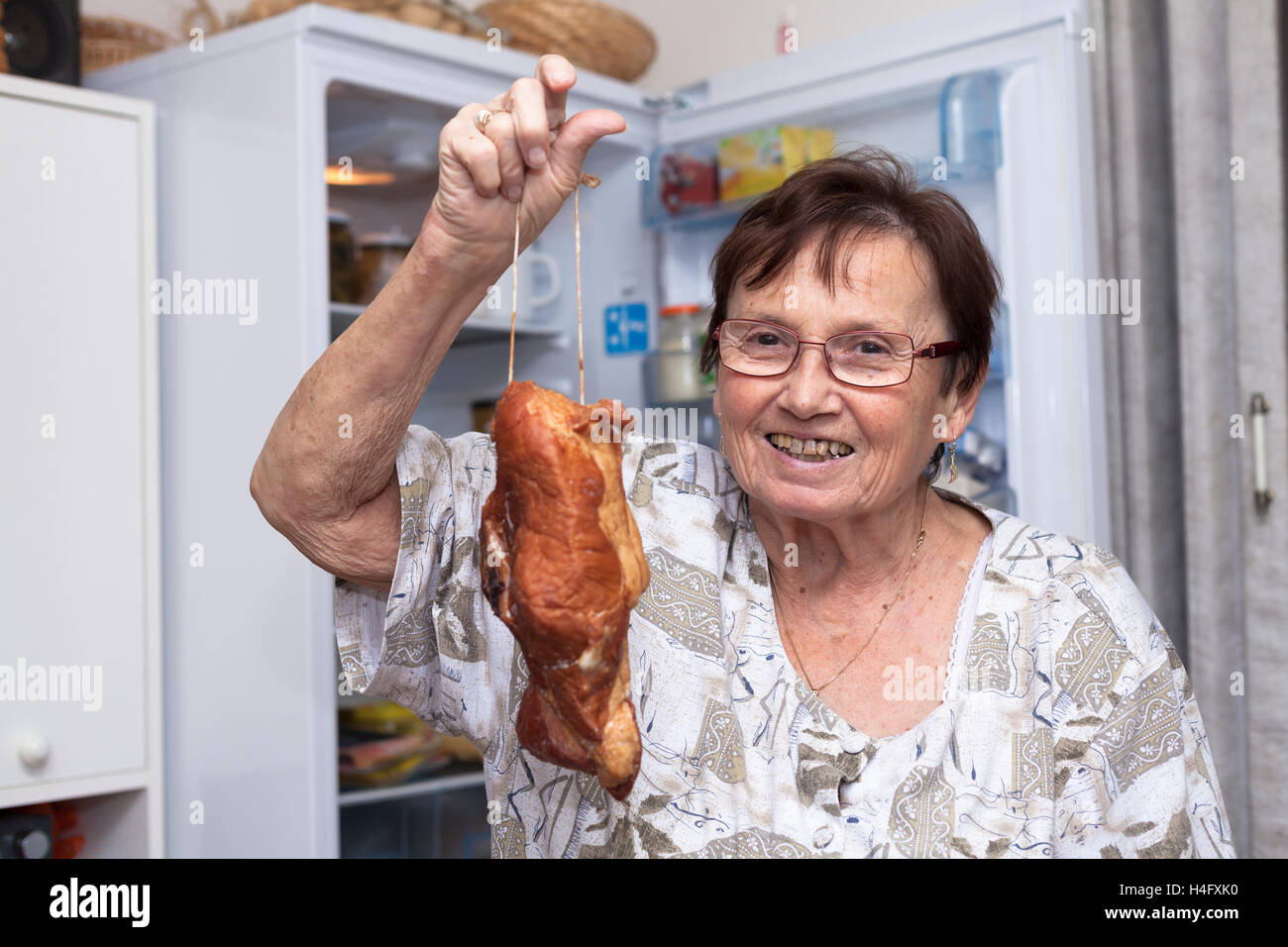 Happy senior woman holding viande fumée alors qu'il se tenait devant l'open frigo dans la cuisine. Banque D'Images