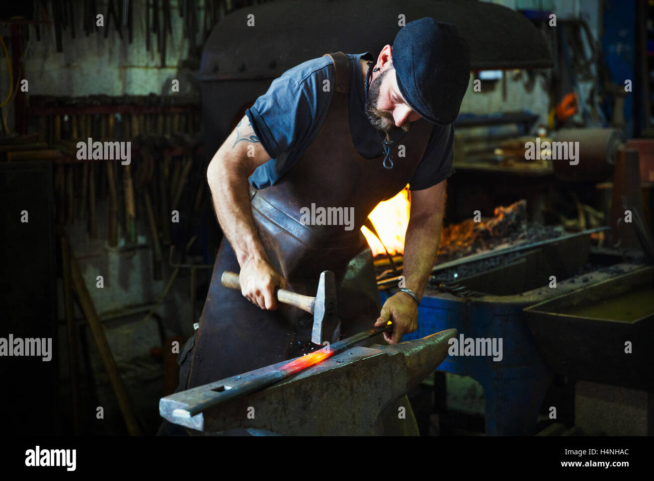 Un forgeron frappe une longueur de métal chaud rouge sur l'enclume avec un marteau dans un atelier. Banque D'Images