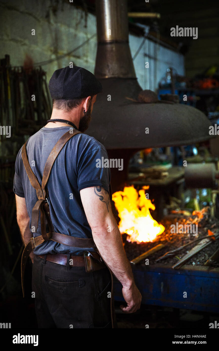Un forgeron portant un tablier de cuir se trouve en face d'un four dans un atelier. Banque D'Images