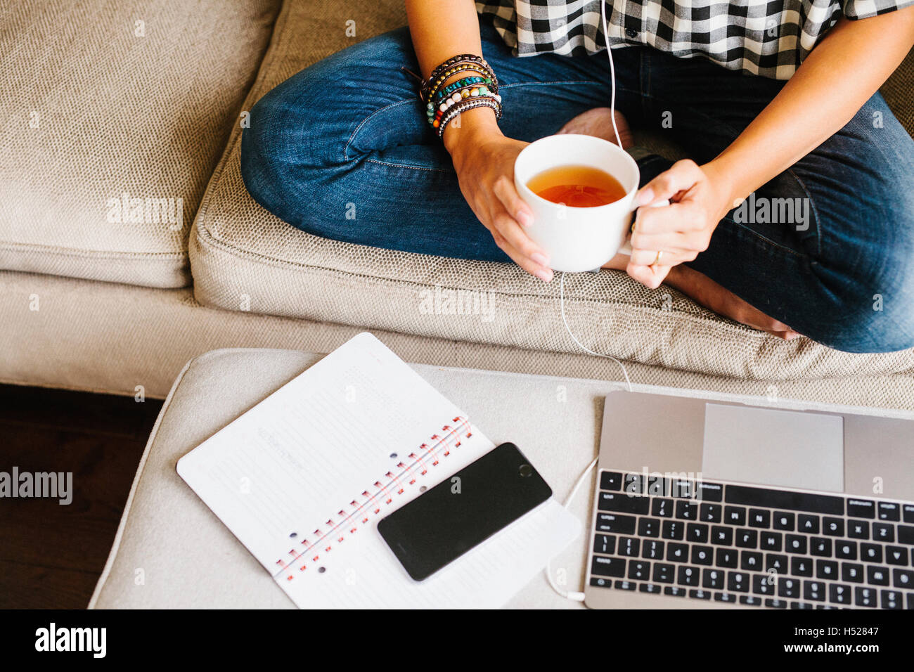 Le port de jeans femme assise sur un canapé en face d'un ordinateur portable, tenant une tasse. Banque D'Images