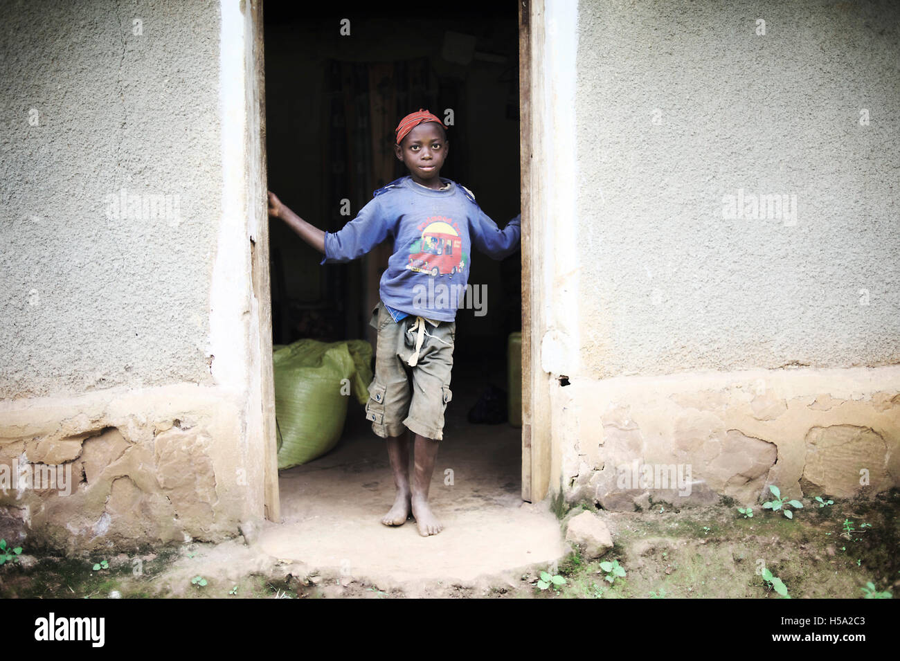 Un jeune enfant se tient à la porte de sa maison ougandais sans chaussures et vêtements débraillés Banque D'Images