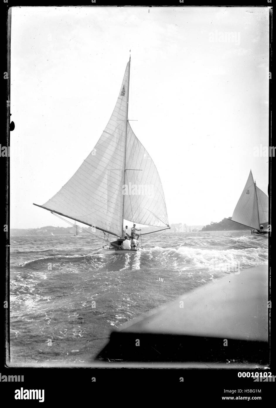 Cette image représente un yacht, peut-être nommé HOANA, naviguant sur le port de Sydney avec cinq membres d'équipage visibles à bord. Le cadre met en valeur le port emblématique et les activités maritimes en Australie. Banque D'Images