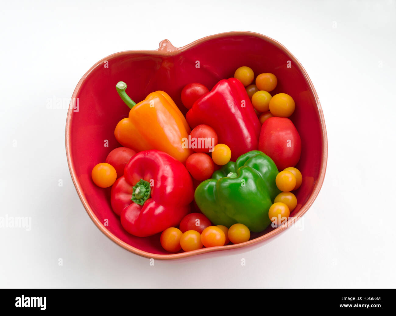 Assortiment de tomates et rouge, jaune et de poivrons verts disposés dans un bol en forme de tomate rouge sur un fond blanc. Banque D'Images