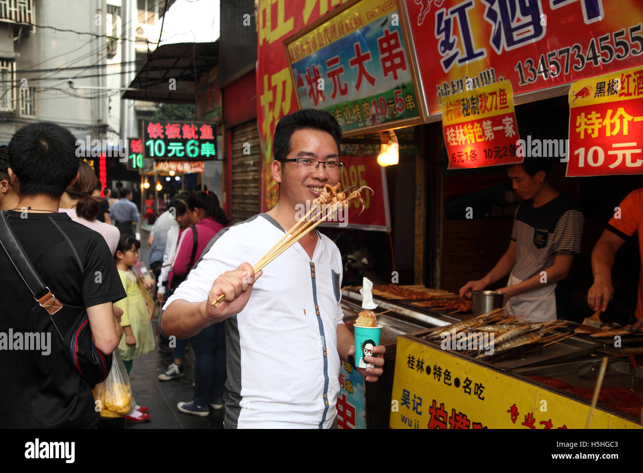 Un jeune Chinois homme sourit tout en mangeant des calmars sur les brochettes, en face d'un stand de restauration rapide. Le marché à Guilin, Chine. 02.05.2 Banque D'Images