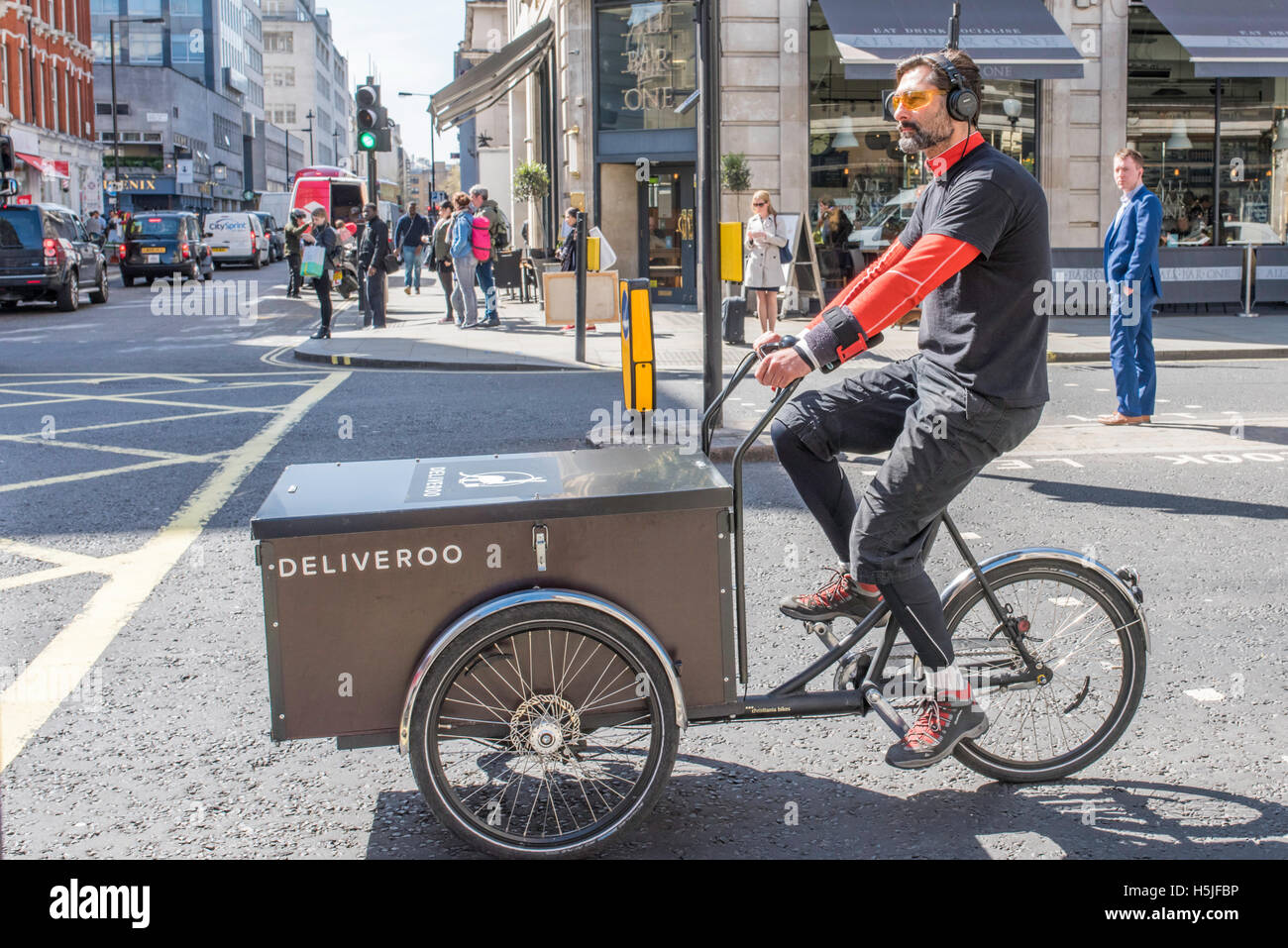 Un cycliste de la livraison des marchandises pour Deliveroo attendant le feu pour changer au vert. Un homme à la recherche sur en arrière-plan. Banque D'Images