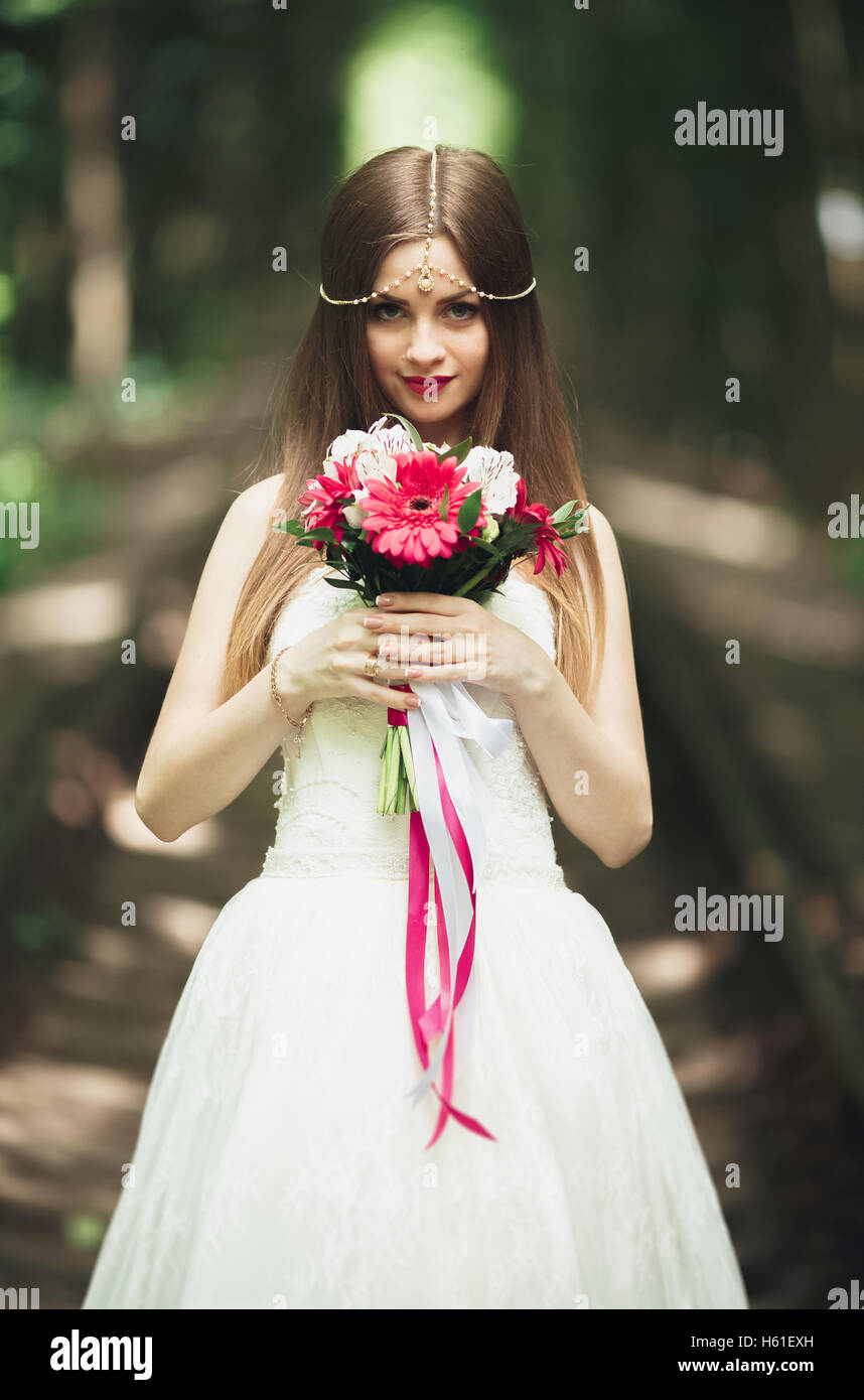 Belle brune mariée élégante en robe blanche holding bouquet posing neat arbres Banque D'Images