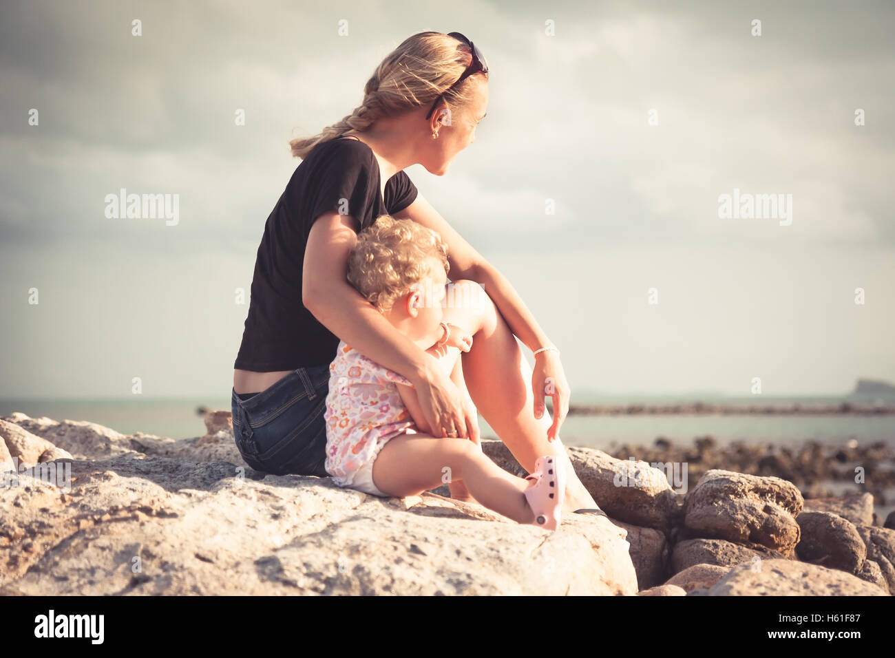 La mère et son bébé assis ensemble sur la plage côte pendant le coucher du soleil et à la recherche dans la distance. Mère et enfant embrasser la lumière du soleil illumine Banque D'Images