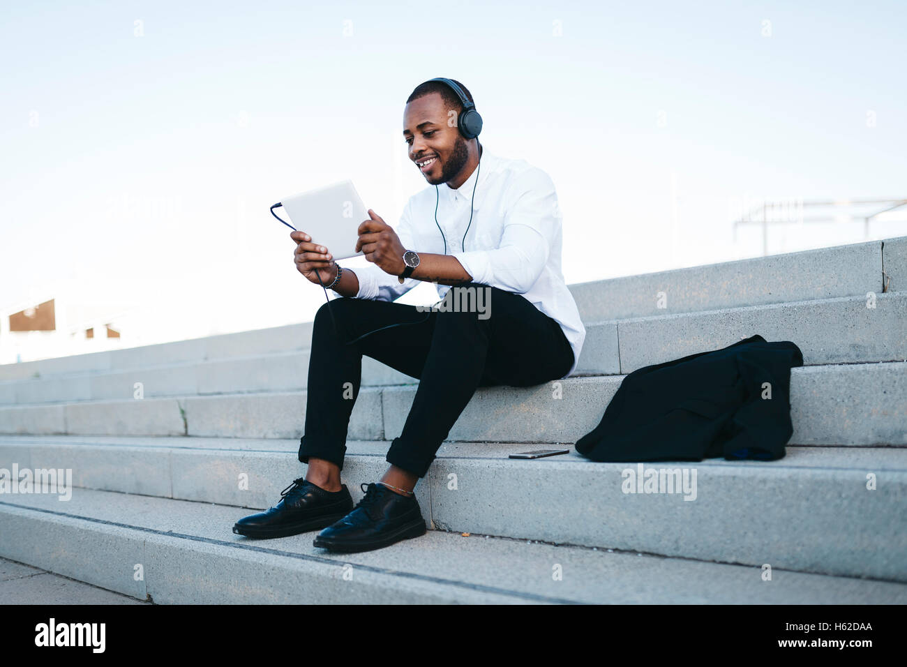 Smiling man sitting on stairs portant des écouteurs à la tablette à Banque D'Images