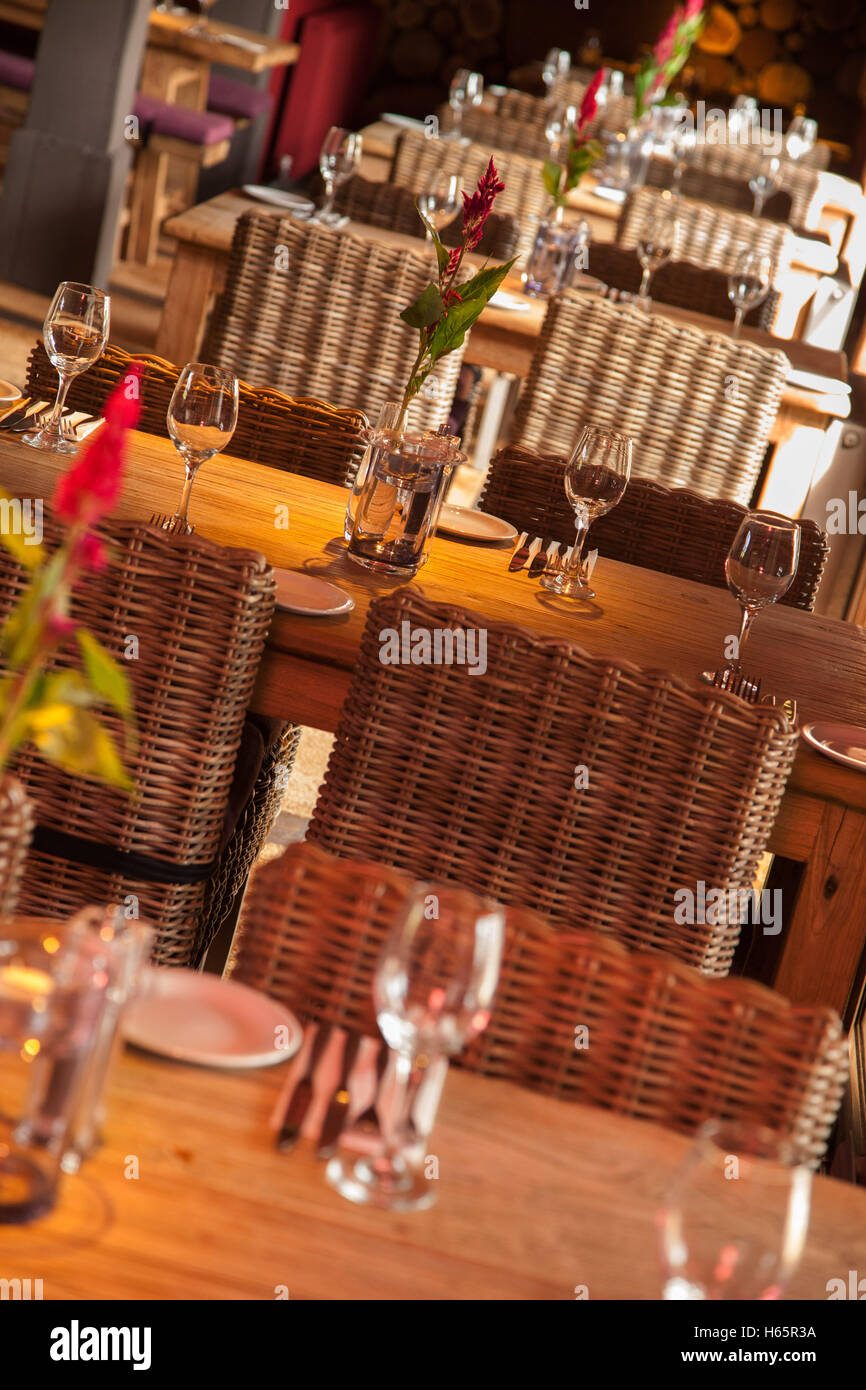 Une rangée de tables en bois avec des chaises en osier posées pour déjeuner dans un restaursant, verres à vin, couverts, fleurs Banque D'Images