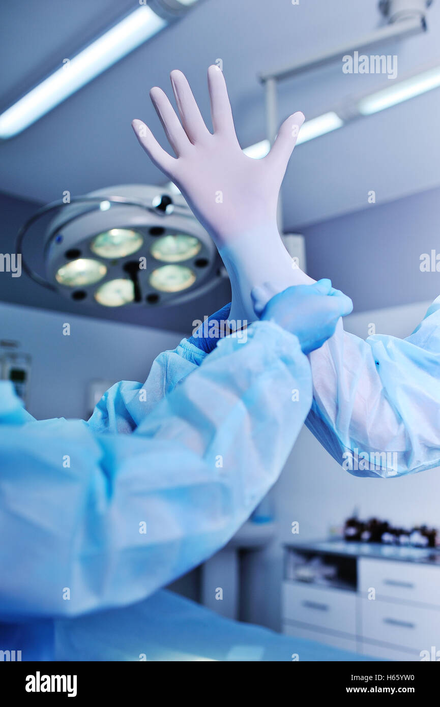 Aide Assistant Le chirurgien mis sur des gants en latex avant l'opération. Une équipe de chirurgiens dans le fond de la lampe d'exploitation prepar Banque D'Images