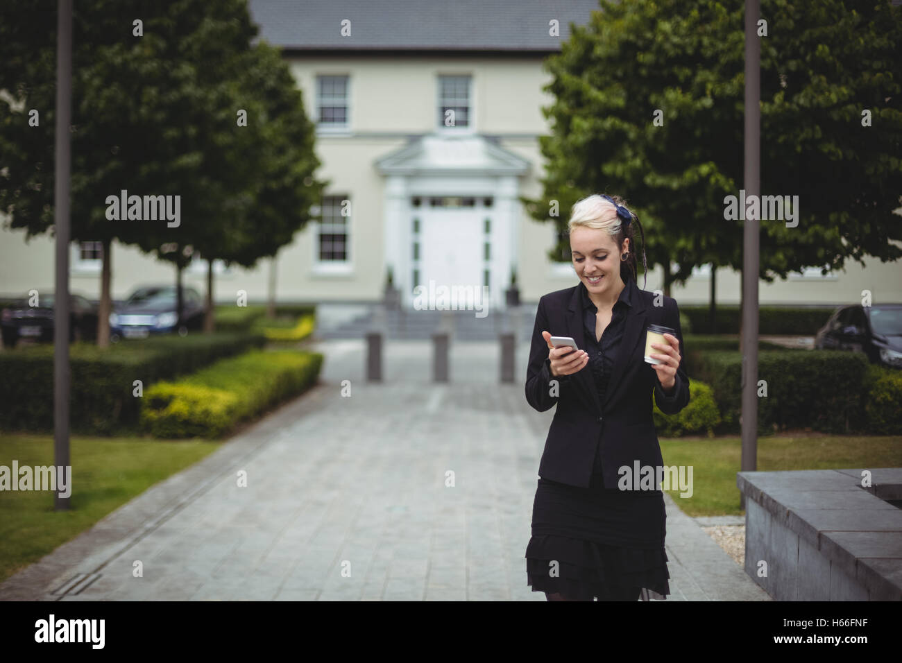 Businesswoman using mobile phone tout en tenant la tasse de café à usage unique Banque D'Images