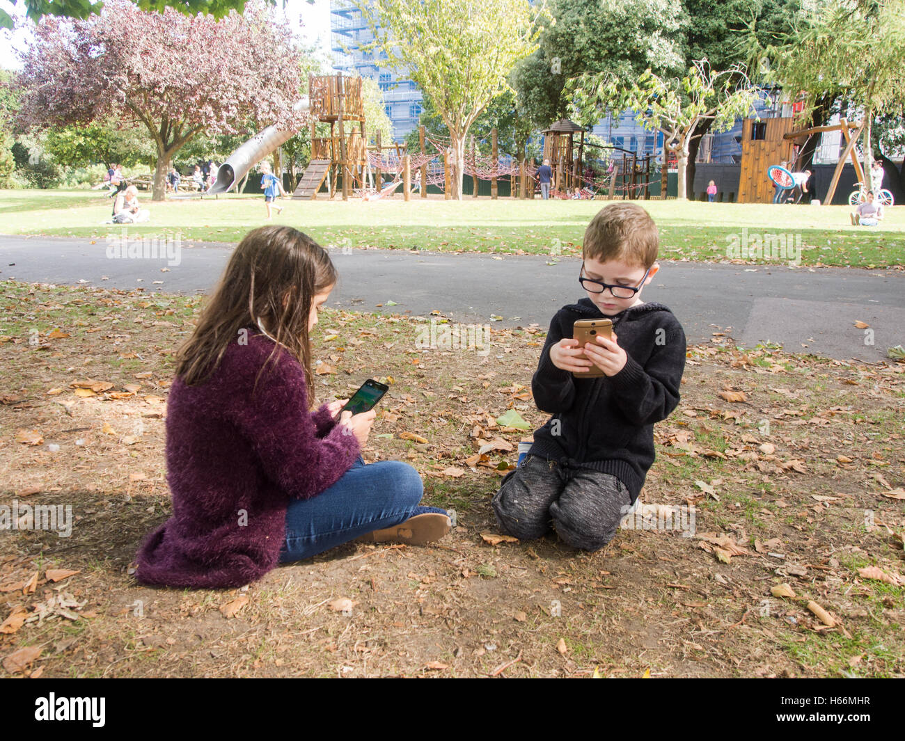 Deux enfants jouent à des jeux sur téléphones mobiles dans un parc public avec une aire de jeux derrière eux. Banque D'Images