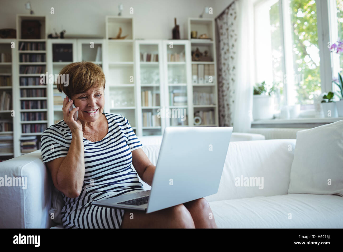 Senior Woman talking on mobile phone while using laptop in living room Banque D'Images