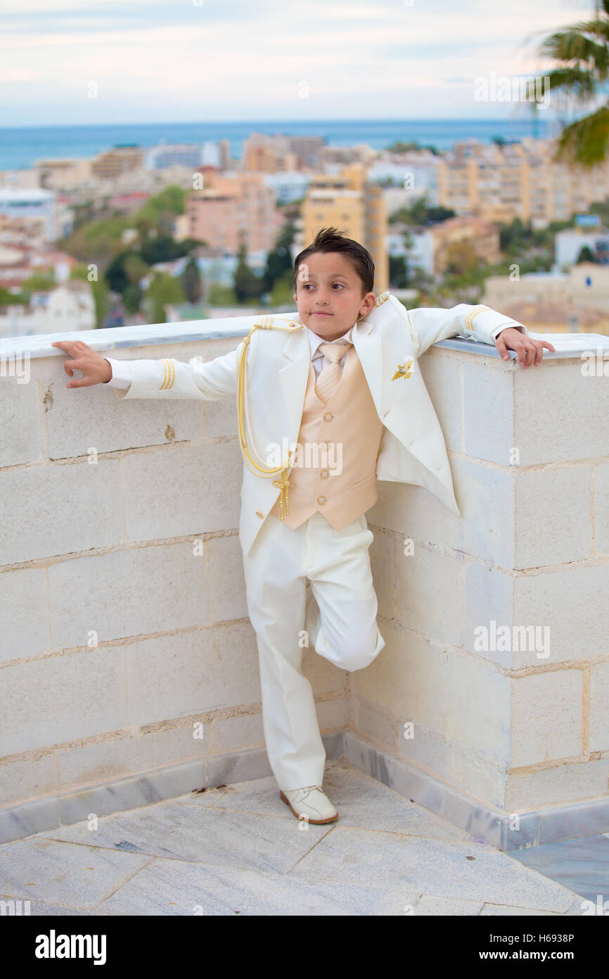 Jeune garçon avec costume blanc s'appuyant sur un mur dans sa Première Communion. Banque D'Images