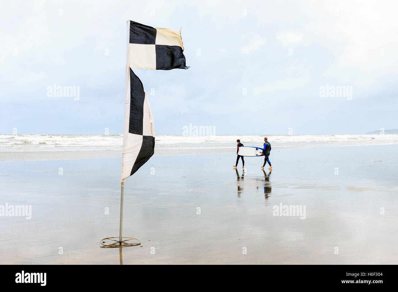 Deux surfers carrying une planche de surf à la mer à marée basse sur la plage à Westward Ho !, Devon, England, UK, un drapeau noir et blanc à l'avant-plan Banque D'Images