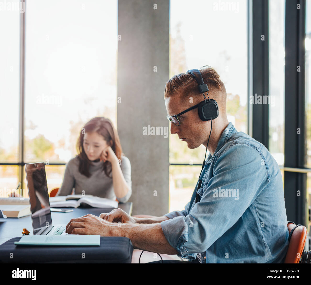 Coup de jeune homme avec un casque à l'aide d'ordinateur portable à la bibliothèque du collège. Les jeunes gens assis à la table fonctionne sur l'affectation d'école Banque D'Images