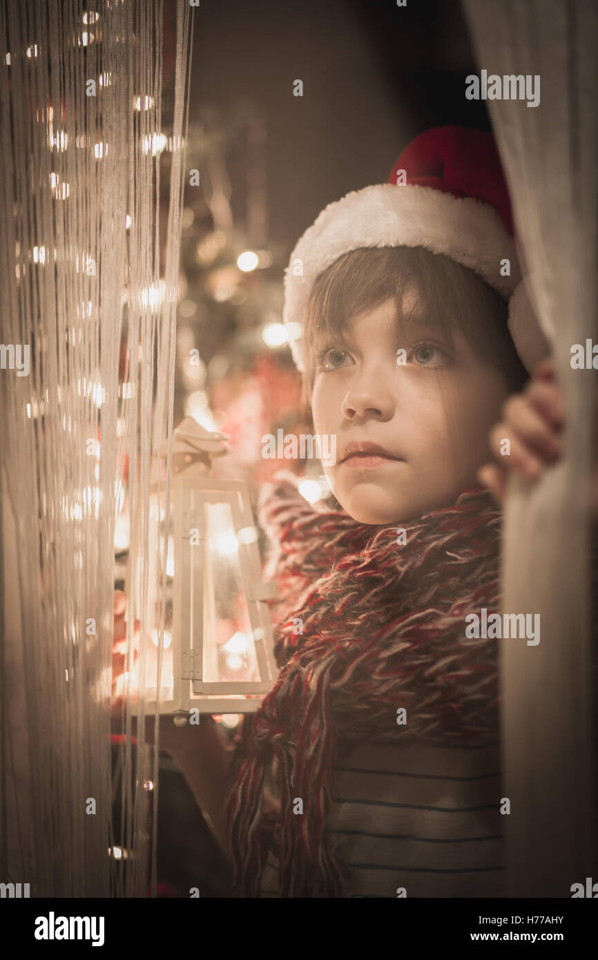 Garçon par une fenêtre wearing christmas santa hat holding une lanterne Banque D'Images