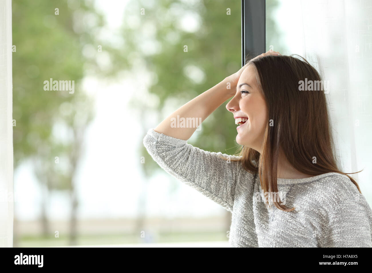 Vue latérale d'un happy girl laughing with hand on head et à l'extérieur à travers une fenêtre à la maison ou chambre d'hôtel Banque D'Images