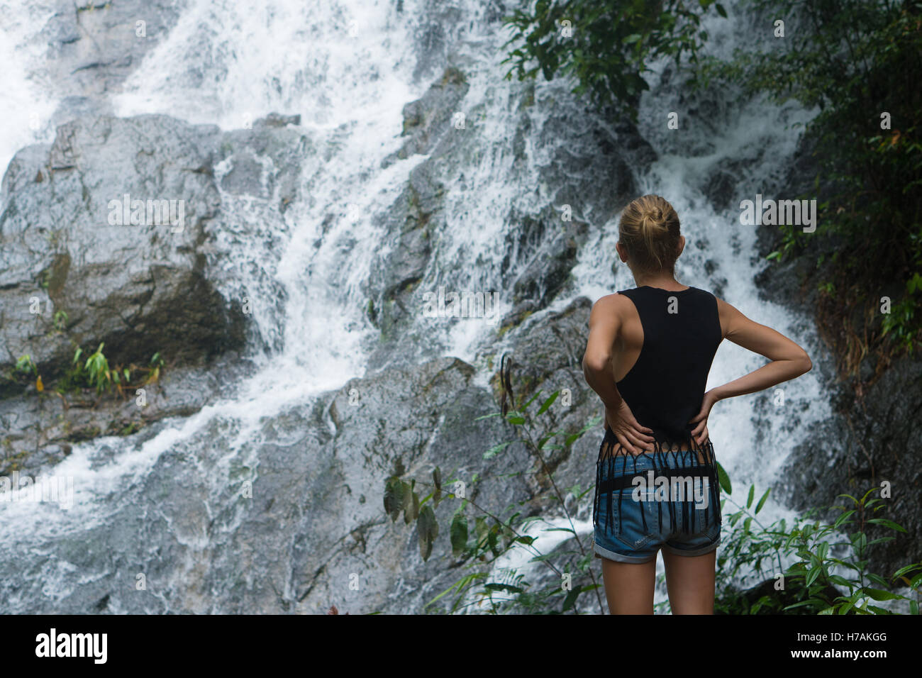 Vue arrière de blonde woman standing on a rock et jusqu'à près de cascade. Bénéficiant d'une femme par une chute d'eau en forêt Banque D'Images