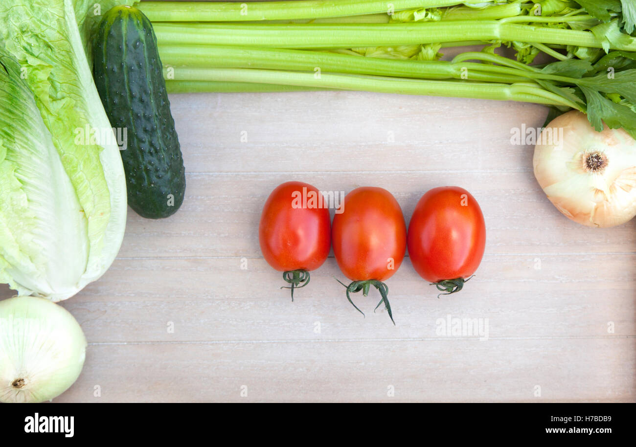 Trois tomates entouré par des légumes frais (l'oignon, le concombre, les feuilles de laitue et de céleri) sur planche à découper en bois Banque D'Images