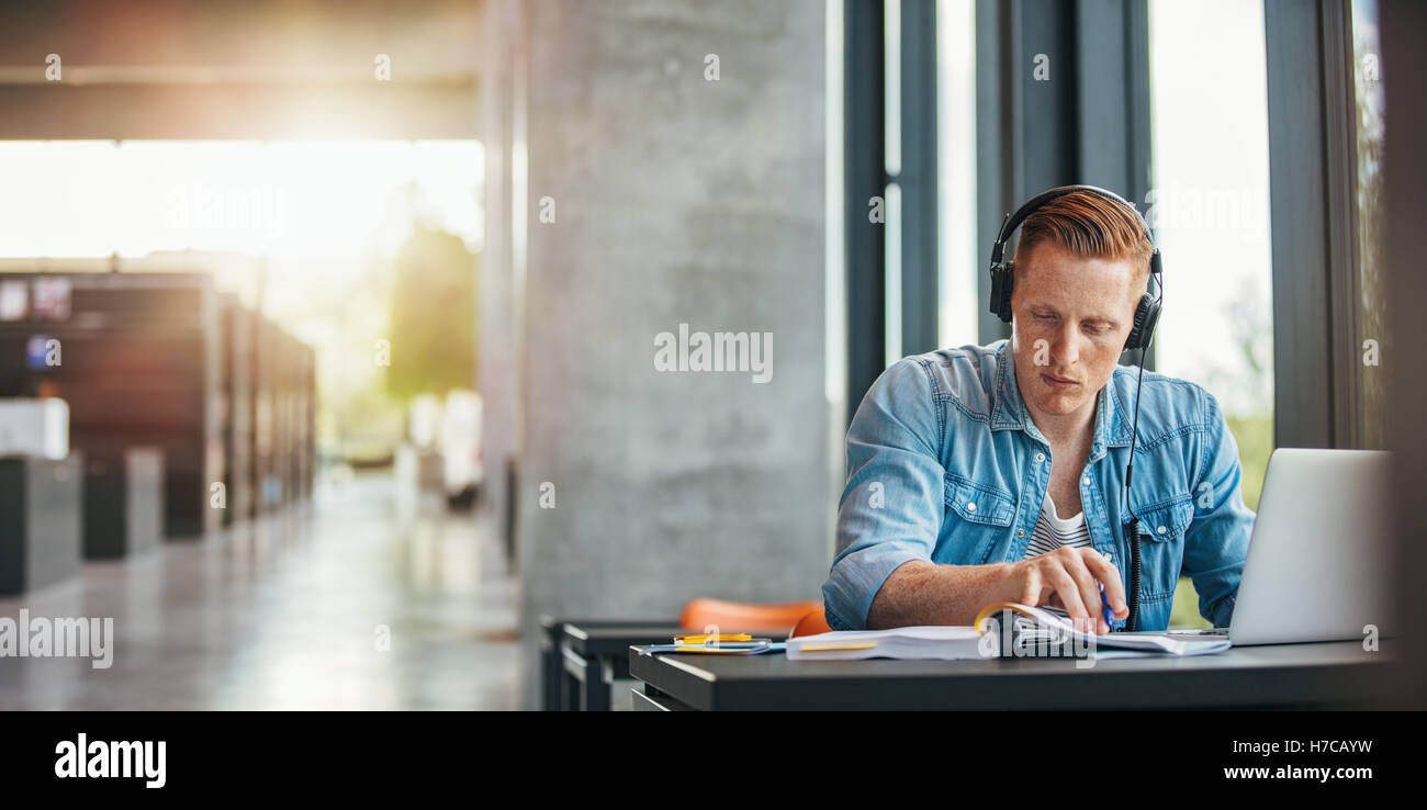 Portrait de jeune étudiant portant des écouteurs à la table dans la bibliothèque et la lecture de livre. Trouver des étudiants de l'Université d'informer Banque D'Images