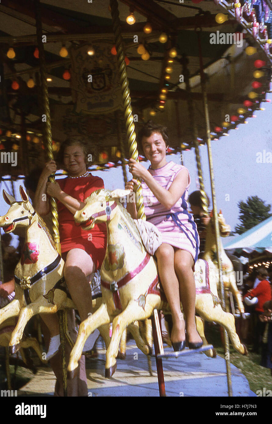 En Angleterre, en 1960, Mère et fille portant des robes équitation sur chevaux de bois sur un champ de foire carrousel avec fille dans une robe rose plus élégamment équitation 'side-selle". Banque D'Images