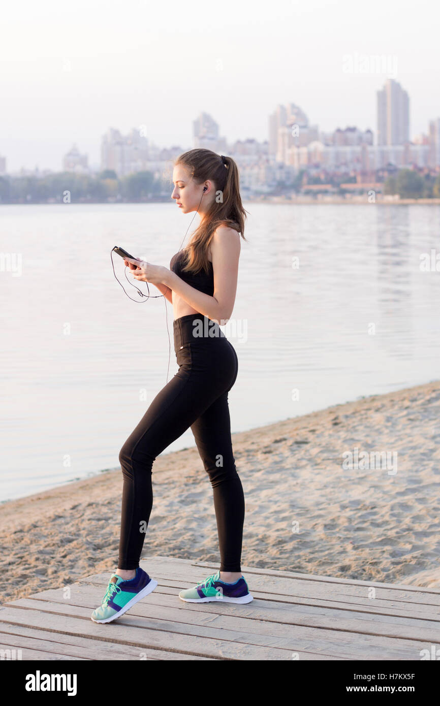 Jolie jeune femme fitness slim est à côte de la rivière dans la matinée à écouter la musique via mobile cell phone player earphon Banque D'Images