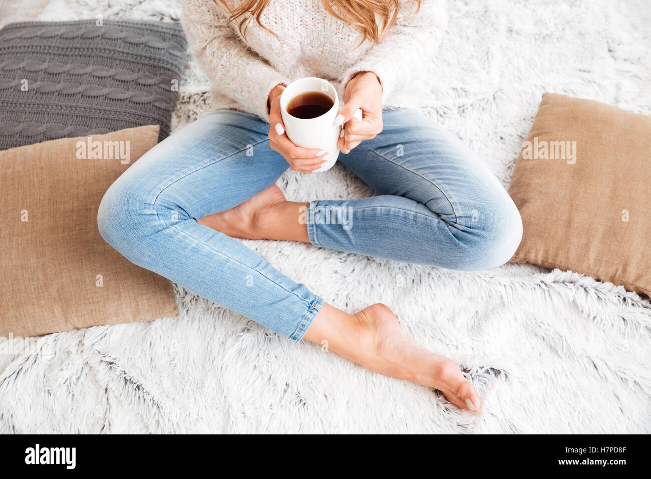 Portrait d'une femme en pull et jeans holding tea cup en position assise avec les jambes croisées à l'intérieur Banque D'Images