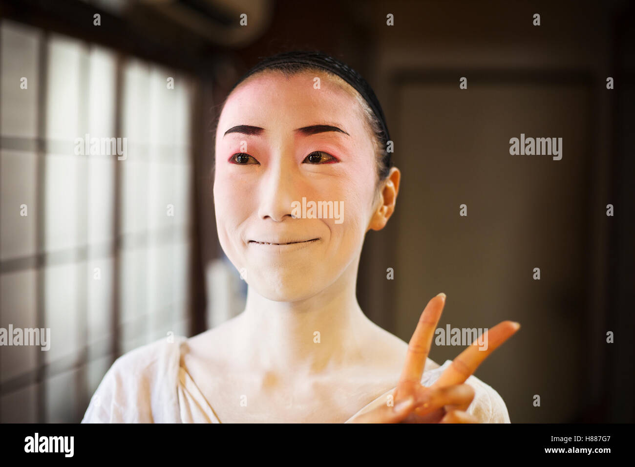 Geisha femme avec un visage blanc traditionnel et maquillage eyeliner lourd holding up deux doigts dans un geste moderne. Banque D'Images