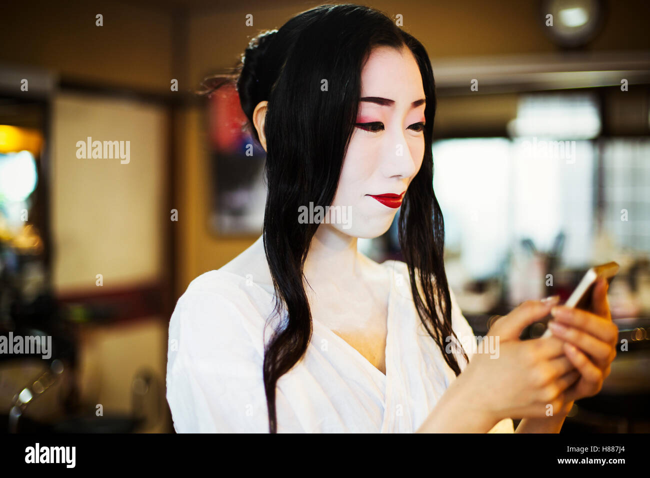 Geisha avec de longs cheveux noirs et un visage blanc traditionnel composent à l'aide d'un téléphone intelligent. Banque D'Images