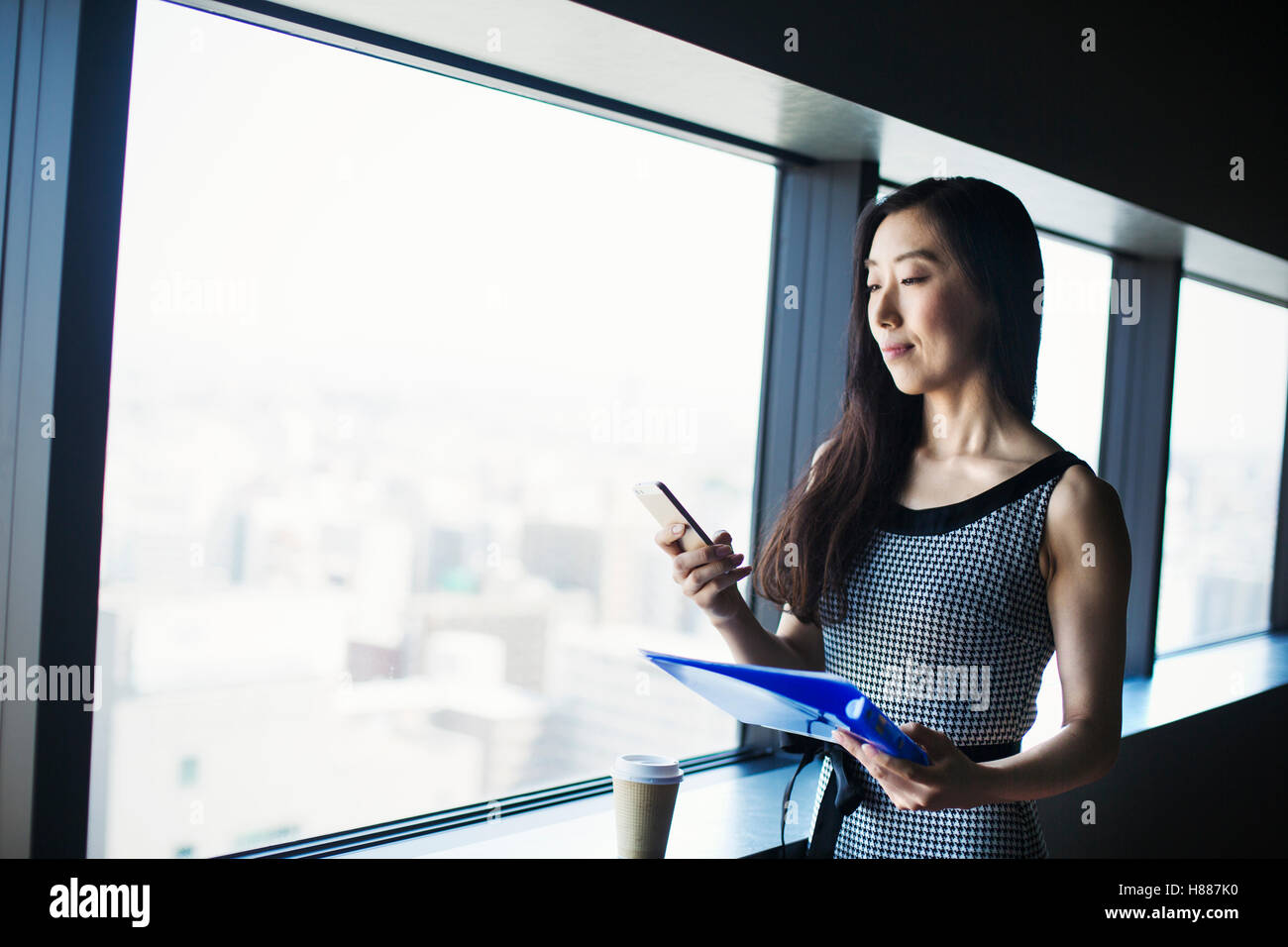 Une femme d'affaires par une fenêtre avec une vue sur la ville, à l'aide de son téléphone intelligent. Banque D'Images Une femme d'affaires par une fenêtre avec une vue sur la ville, à l'aide de son téléphone intelligent. Banque D'Images