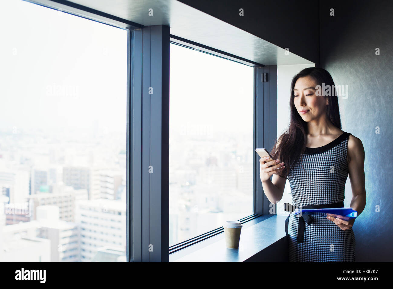 Une femme d'affaires par une fenêtre avec une vue sur la ville, à l'aide de son téléphone intelligent. Banque D'Images Une femme d'affaires par une fenêtre avec une vue sur la ville, à l'aide de son téléphone intelligent. Banque D'Images