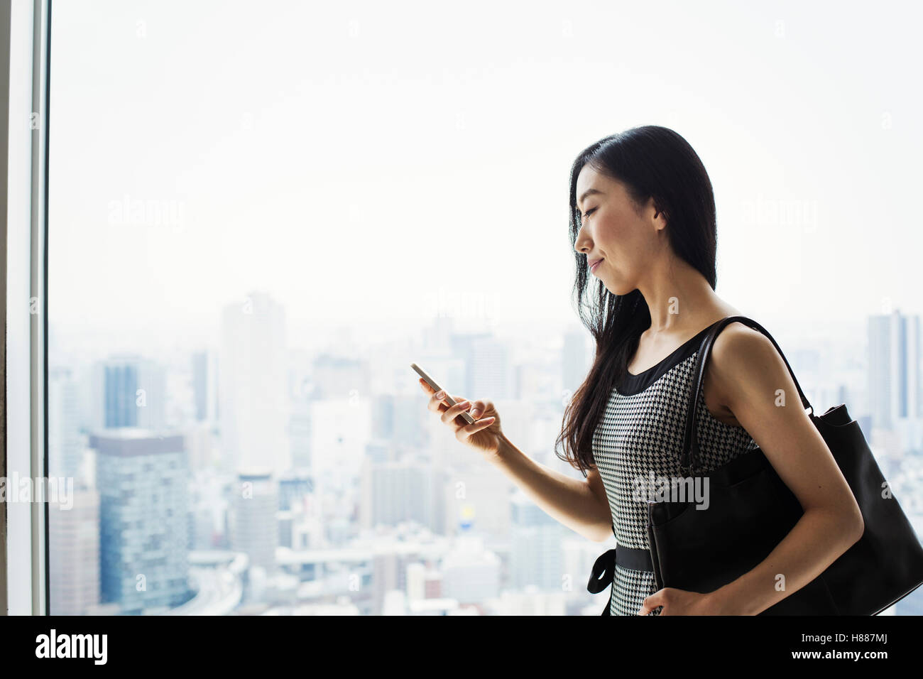 Une femme d'affaires par une fenêtre avec une vue sur la ville, à l'aide de son téléphone intelligent. Banque D'Images Une femme d'affaires par une fenêtre avec une vue sur la ville, à l'aide de son téléphone intelligent. Banque D'Images