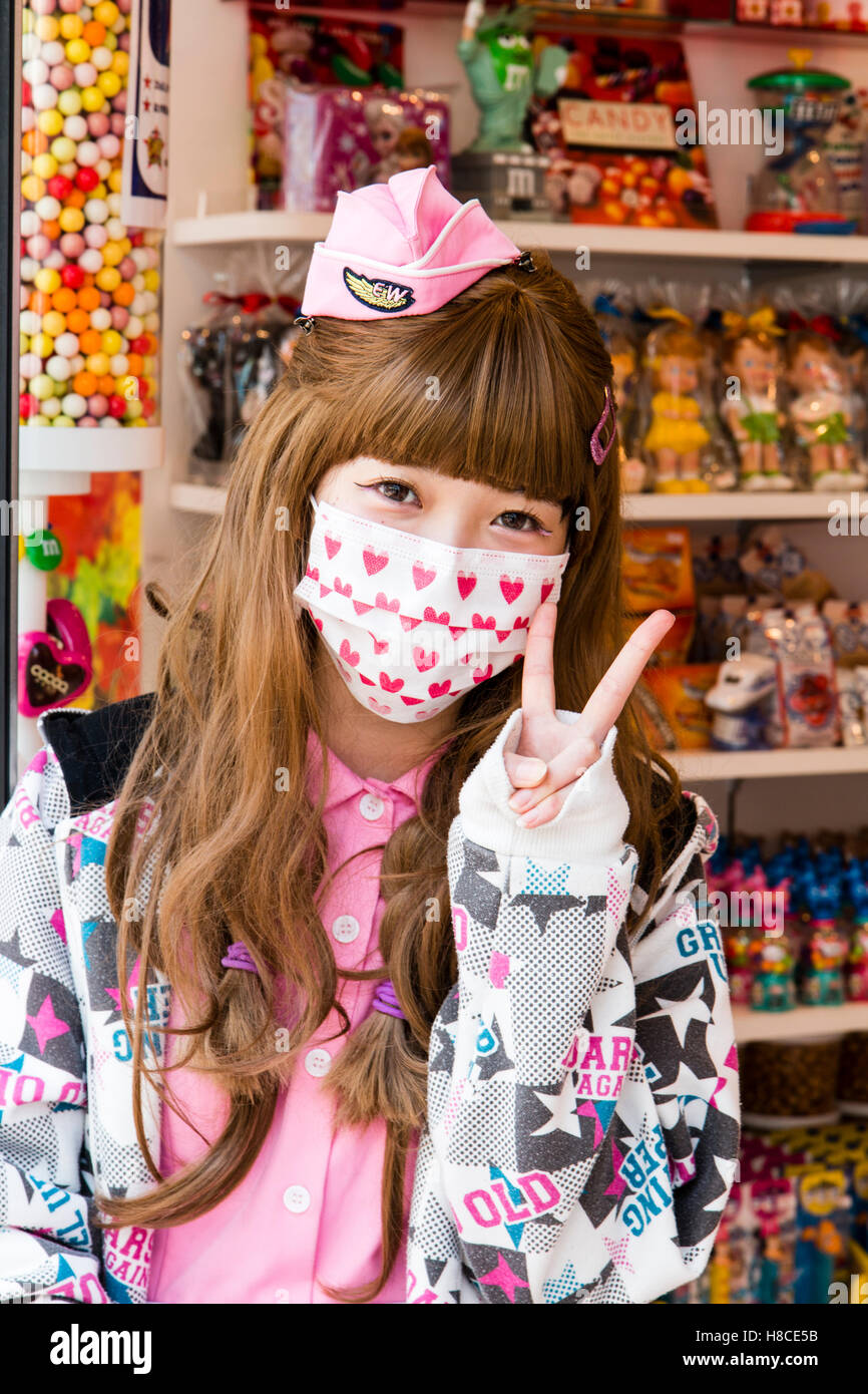 Japon, Tokyo, Harajuku, Takeshita-dori. Les adolescents japonais femme shop assistant, posant pour viewer avec mini rose airline hat masque de visage et signe de la paix Banque D'Images