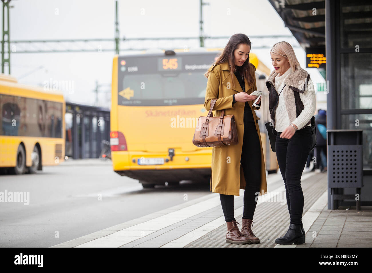 Deux jeunes femmes se tenant debout avec les smartphones à la station de bus Banque D'Images